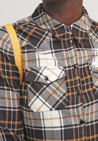 Camisa de franela a cuadros en marrón, blanco y negro con dos bolsillos delanteros y cuello abotonado. Correa amarilla visible.