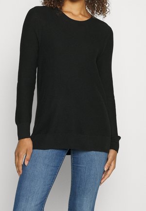 Femme portant un pull noir à manches longues et un jean bleu, debout devant un fond blanc uni.