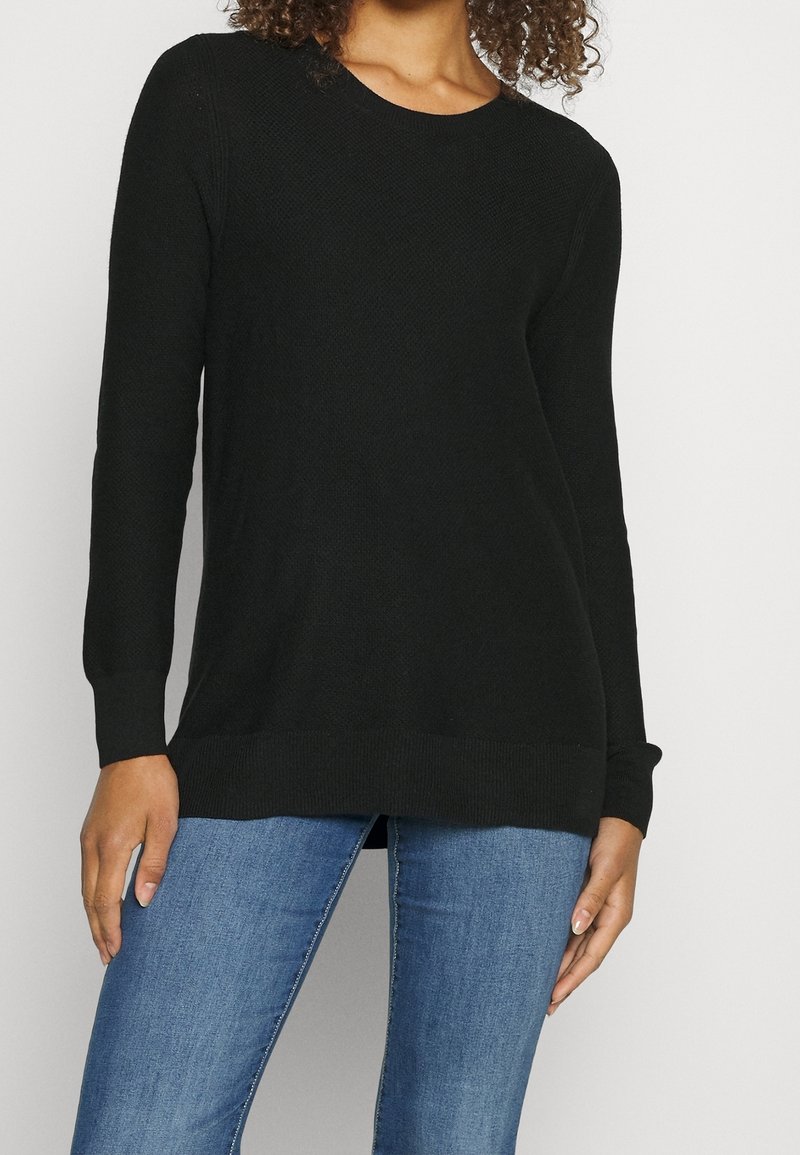 Femme portant un pull noir à manches longues et un jean bleu, debout devant un fond blanc uni.