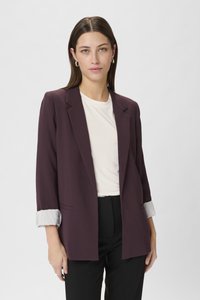 Blazer marrone con una vestibilità comoda, revers a punta e polsini a righe arrotolati. Indossato sopra un top bianco, abbinato a pantaloni neri.