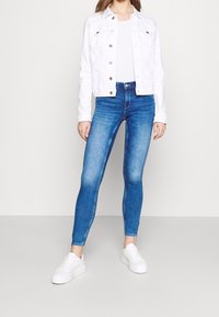 Žena v bílém džínovém saku, bílém topu, modrých skinny džínách a bílých platformních teniskách stojí na jednobarevném pozadí.