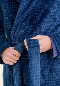 Manos atando el cinturón de un albornoz texturizado de color azul oscuro sobre una camisa de manga larga con un puño azul visible.