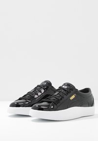 Sneakers in pelle verniciata nera con suole in gomma bianca, lacci neri e dettagli del logo dorati. Punta rotonda e design elegante con dettagli texture.