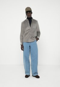 Camisa de pana gris con cuello abotonado, bolsillo frontal; combinada con unos jeans de mezclilla azul claro sueltos y una gorra de cuadros. Zapatos marrones.