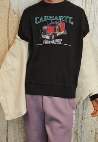 Sudadera negra con un gráfico de camión, cuello y puños acanalados, combinada con un exterior beige, complementada con pantalones de chándal de un tono púrpura claro.
