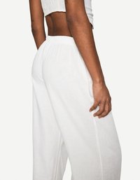 Personne à la peau marron portant un haut blanc court et un pantalon blanc fluide, debout, mettant l'accent sur le dos et le profil latéral de la tenue.