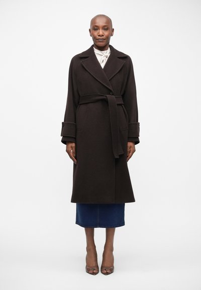 Brown wool coat with a double-breasted design, wide lapels, a belted waist, and side pockets. Worn over a blue denim skirt and heels.