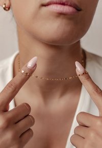 Złoty metalowy naszyjnik z owalnymi ogniwami, trzymany przez dwa palce, w komplecie z długimi różowymi paznokciami. Żadne inne akcesoria nie są widoczne.