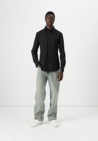 Svart langermet skjorte med knappestolpe og brystlomme, sammen med lyseblå, løstsittende jeans og hvite joggesko.