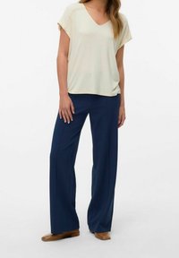 Kermanvärinen v-mallinen toppi, jossa on lyhyet hihat, yhdistettynä tummansinisiin leveälahkeisiin housuihin. Asun täydentävät ruskeat tasapohjaiset kengät, ja näkyvissä on sileät kangaspinnat.