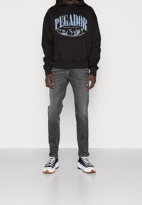 Schwarzer Hoodie mit blauer "PEGADOR" Grafik und Palmen-Design, kombiniert mit grauen Jeans und schwarz-weißen Sneakern mit orangefarbenen Akzenten.