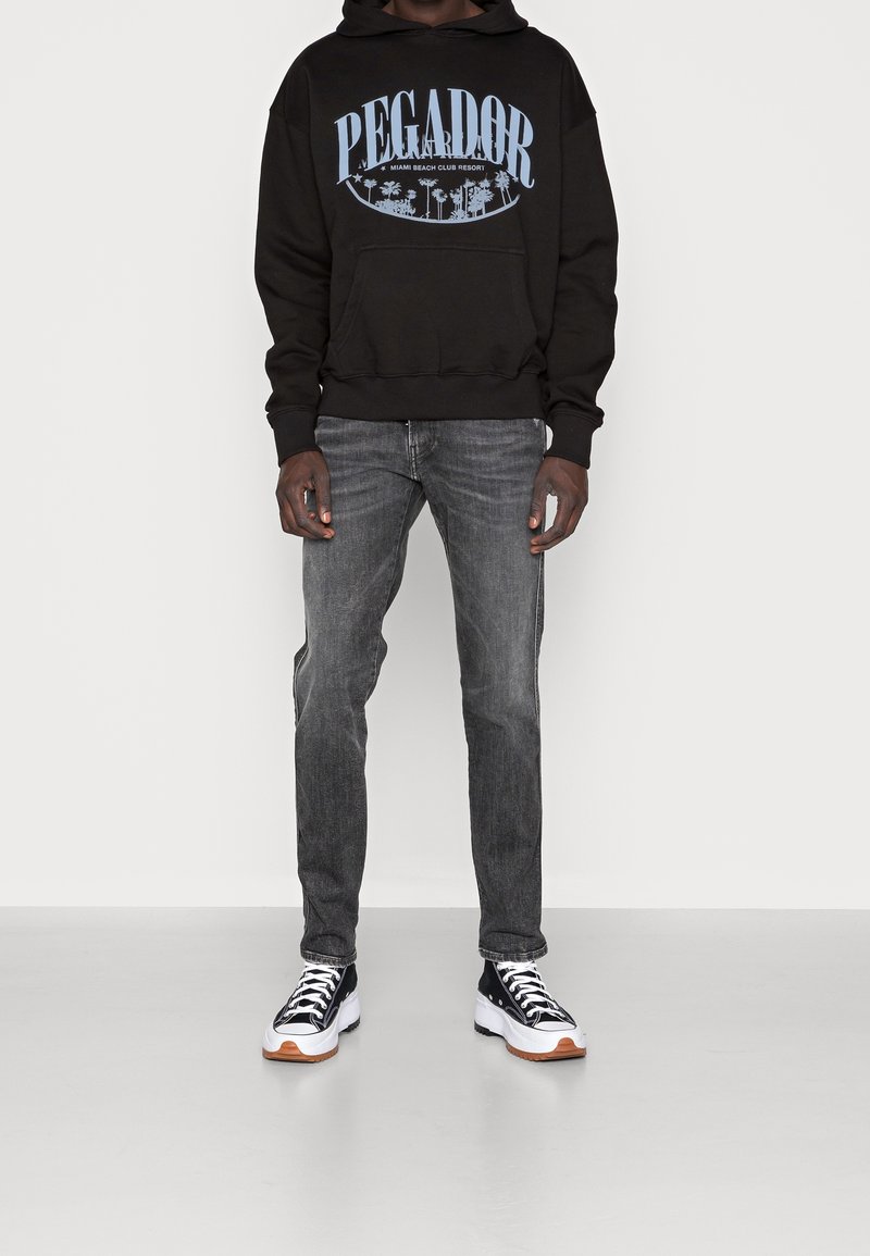 Schwarzer Hoodie mit blauer "PEGADOR" Grafik und Palmen-Design, kombiniert mit grauen Jeans und schwarz-weißen Sneakern mit orangefarbenen Akzenten.