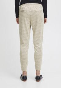 Karierte Hose mit elastischem Bund, schmaler Schnitt, zwei Rückentaschen und einem creme- und schwarzfarbenen Muster. Getragen mit schwarzen Schuhen.