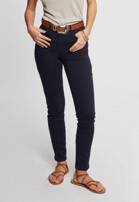 Pantalons ajustés bleu marine avec un léger stretch, associés à une ceinture en cuir marron avec une boucle en argent, portés avec des sandales strappy marron.