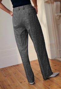 Persona che indossa pantaloni grigi a vita alta e a gamba larga, texurizzati, e décolleté beige a punta, in piedi su un pavimento di legno vicino a una parete bianca e un camino.