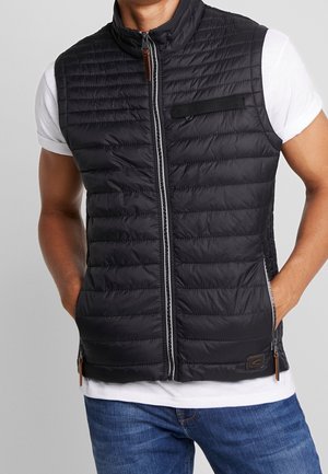 Mężczyzna ubrany w czarną pikowaną kamizelkę bez rękawów na białej koszulce, z rękami w kieszeniach kamizelki, na jednolitym tle.