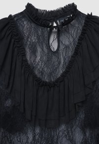 Blouse noire en tissu dentelle, col et manches volantés, avec une ouverture en forme de trou de serrure au niveau du cou. Aspect texturé et transparent.