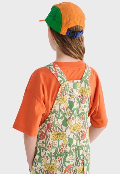 Enfant portant un t-shirt orange et une salopette à motifs floraux, avec une casquette orange et verte, dos tourné, sur un fond clair.