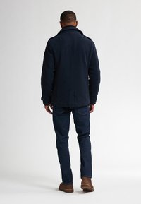 Cappotto navale con alto colletto e dettagli con bottoni, indossato sopra jeans blu scuro e stivali marroni, visto da dietro. Texture liscia.