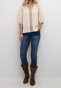 Blouse en lin beige clair avec un décolleté à lacets, des manches bouffantes trois-quarts et des garnitures en dentelle décoratives sur le devant, associée à un jean bleu et des bottes marron.