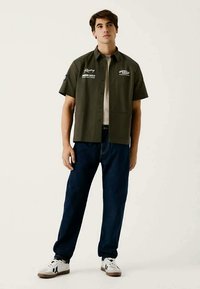 Camisa verde-oliva de manga curta com logótipos bordados, fecho de botão, combinada com umas calças de ganga azuis largas e ténis brancos.