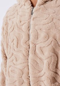 Giacca con cappuccio beige, con zip, realizzata in un morbido tessuto testurizzato, caratterizzata da un motivo ondulato sulla superficie e una zip argentata.
