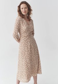 Beiges Polka-Dot-Kleid mit V-Ausschnitt, dreiviertellangen Ärmeln, gerafftem Taillenbund und fließendem Saum. Hergestellt aus leichtem Stoff.