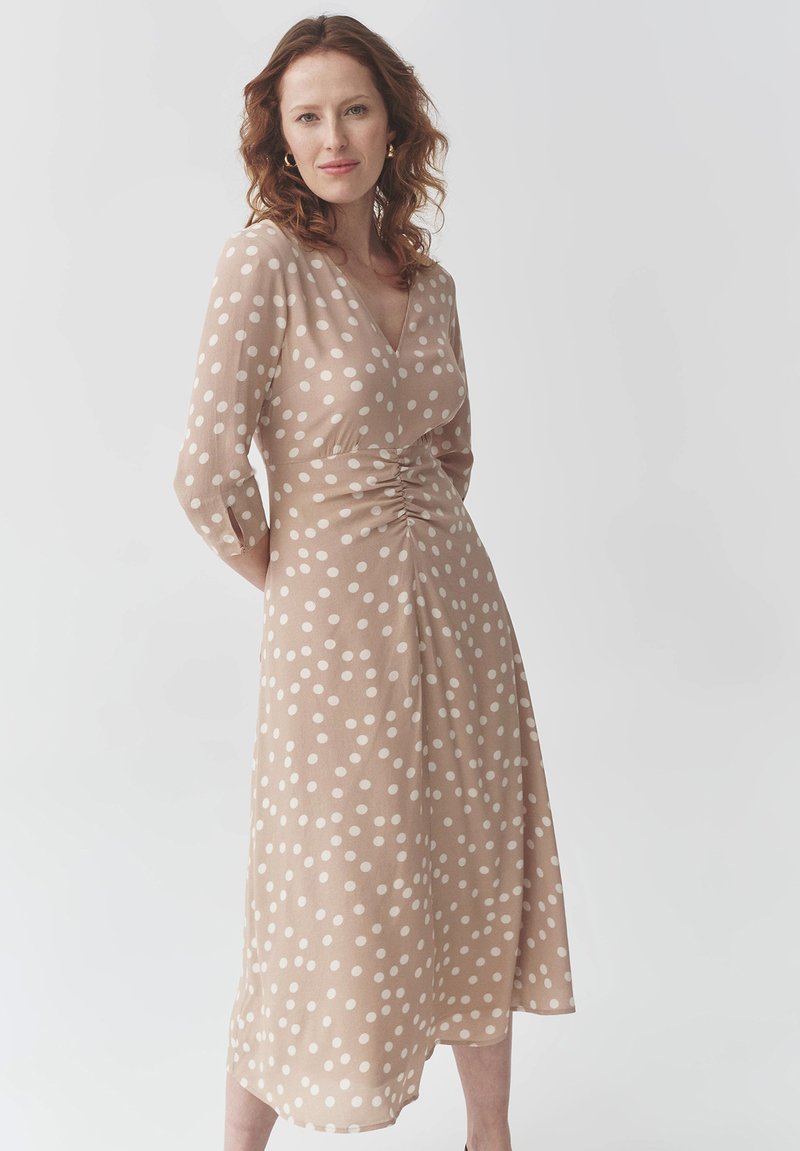 Beiges Polka-Dot-Kleid mit V-Ausschnitt, dreiviertellangen Ärmeln, gerafftem Taillenbund und fließendem Saum. Hergestellt aus leichtem Stoff.