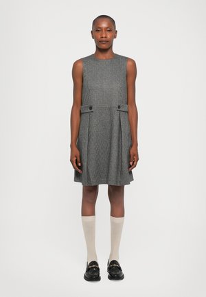 Donna in piedi contro uno sfondo neutro, che indossa un vestito grigio senza maniche, calze beige al ginocchio e mocassini neri con dettagli dorati.
