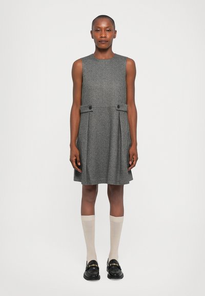 Donna in piedi contro uno sfondo neutro, che indossa un vestito grigio senza maniche, calze beige al ginocchio e mocassini neri con dettagli dorati.