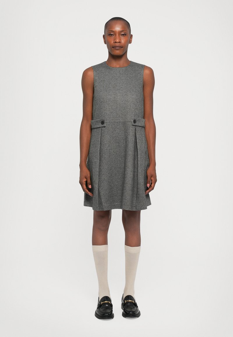 Frau, die vor einem schlichten Hintergrund steht und ein ärmelloses graues Kleid, beiges kniehohes Socken und schwarze Slipper mit goldenen Details trägt.