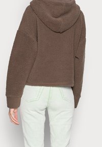 Brauner kurz geschnittener Fleece-Hoodie mit strukturierter, weicher Oberfläche und Kapuze; kombiniert mit mintgrünen Cordhosen mit vertikalen Rillen.