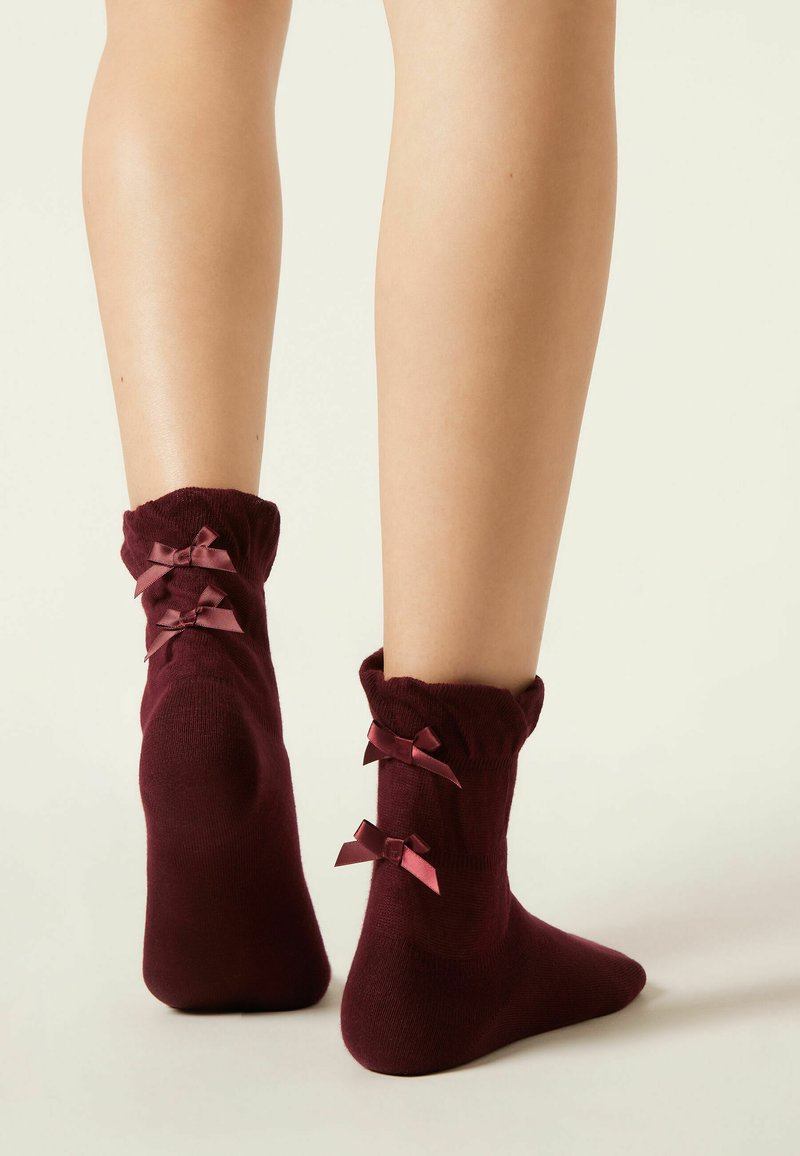 Burgundy ankelsockor med rynkade kant och två små satängrosetter på baksidan. Mjuk textur och åtsittande design.