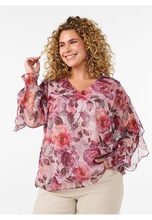 Lächelnde Frau mit lockigem Haar trägt eine rosa, durchsichtige Bluse mit Blumenmuster und langen Puffärmeln sowie hellfarbige Hosen.