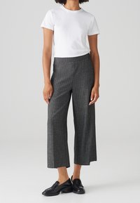 Femme portant une chemise blanche à manches courtes, un pantalon gris ample retroussé et des mocassins noirs, debout devant un fond clair uni.