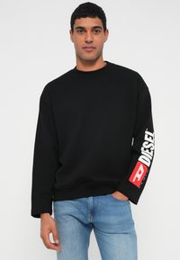 Musta pusero, jossa on pyöreä kaula-aukko, laskettu olkapää ja pitkät hihat. Vasemmassa hihassa on valkoinen logo. Päällä siniset farkut.