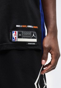 Κοντινό πλάνο μαύρης φανέλας και σορτς μπάσκετ με το κείμενο "Μια φορά Knick, πάντα Knick," ετικέτα μεγέθους 44 και ένα χέρι που ακουμπά δίπλα στο ύφασμα.