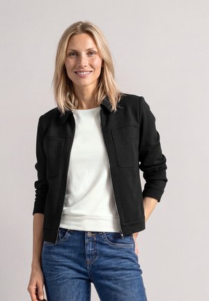 Une femme souriante porte une veste noire zippée sur le devant, par-dessus un haut blanc et un jean bleu, sur un fond clair et uni.
