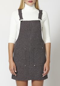 Svartvitt rutigt hängselklänning med framfickor, justerbara metallremmar och rak skärning, bärs över en åtsittande vit polotröja.