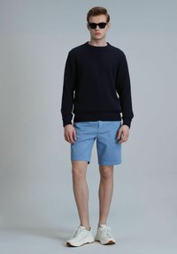 Marineblauwe geribbelde gebreide trui, lichtblauwe shorts en witte sneakers met bruine accenten. Model draagt zwarte zonnebril en staat tegen een lichte achtergrond.