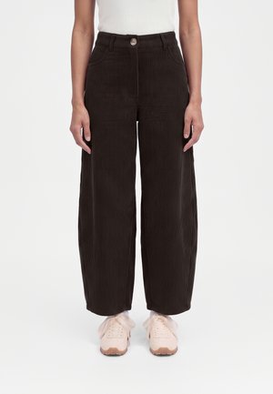 Personne portant un pantalon large en velours côtelé marron foncé taille haute et des baskets roses claires avec des lacets ruban, debout devant un fond blanc.