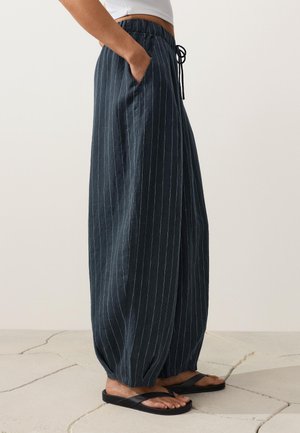 Personne portant un pantalon ample bleu foncé à fines rayures avec poignets élastiques, taille élastique avec cordon de serrage, poche latérale et tongs noires.