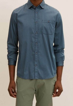 Mann trägt blau gemustertes langärmliges Hemd und olivgrüne Hose, steht vor einem einfarbigen hellen Hintergrund.