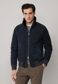 Mornarica suknja od antilopa s rebrastim manžetama i ovratnikom, s prednjim zatvaračem i bočnim džepovima, nosi se preko svijetlosive džemperice.