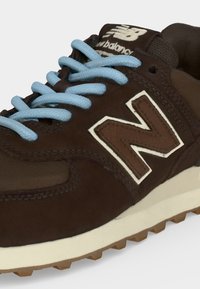 Καφέ αθλητικό παπούτσι New Balance με γαλάζια κορδόνια, λευκή ενδιάμεση σόλα και υφασμένη εξωτερική σόλα από καουτσούκ, παρουσιάζοντας πλευρικές και επάνω λεπτομέρειες.