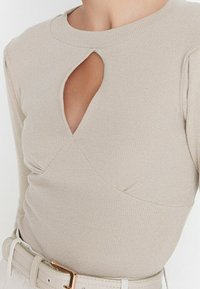 Haut beige à manches longues côtelées avec un col rond et une ouverture en forme de goutte, orné de détails en tissu froncé à la taille.