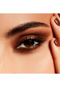 Gros plan sur un œil avec un fard à paupières marron foncé et doré, des cils longs et des sourcils bien définis, arborant un teint naturel et un vernis à ongles marron.