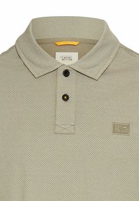 Beige polo skjorte med et struktureret mønster, tre-knaps placket og et lille broderet logo på venstre bryst. Boligstof i bomuldsblanding.