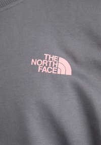 Hanorac din țesătură gri, cu o textură ribbed și un logo roz al "The North Face" pe piept.
