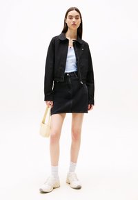 Sort kort jakke over en lyseblå top, kombineret med et sort denimskørt. Tilbehør inkluderer en lille beige taske og hvide sneakers med grå detaljer.